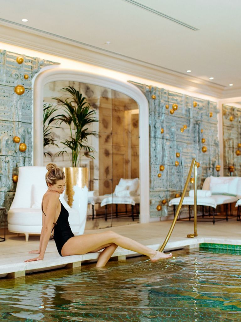 A woman in a black swimsuit dipping her feet  into an indoor pool.